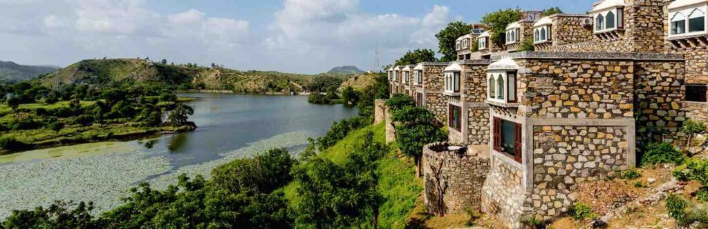 Stone buildings overlook a lush hillside and a tranquil lake, surrounded by green hills under a partly cloudy sky—a scene reminiscent of the view from the best rooftop restaurants in Udaipur.