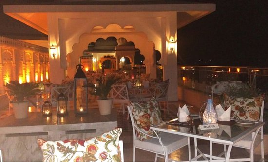 Rooftop dining area at night with ornate decor, floral upholstery, and soft lighting, overlooking a cityscape.