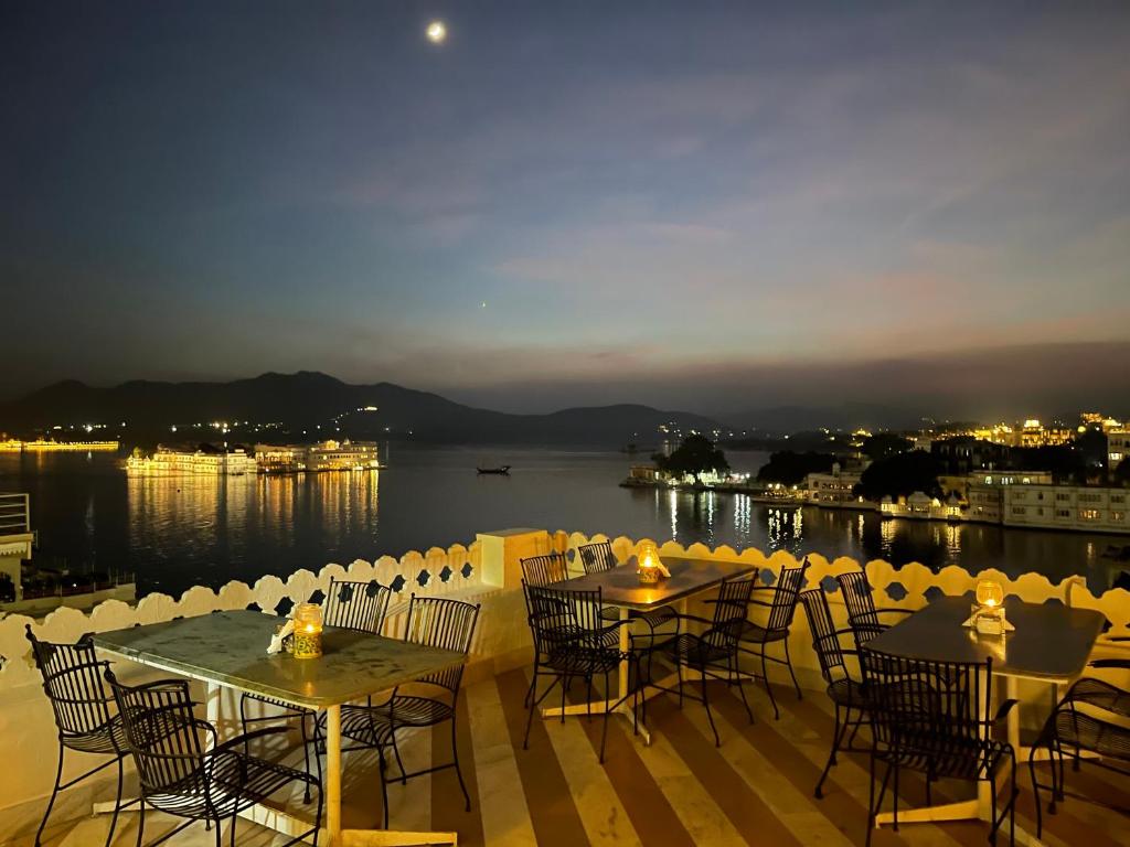 Terrace view at dusk with patio furniture overlooking a serene lake, distant mountains, lit-up cityscape, and a visible crescent moon in the sky.