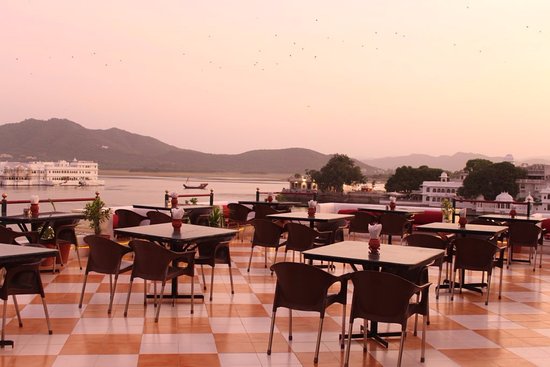 Rooftop restaurant at twilight overlooking a lake with distant hills and boats, featuring several dining tables and chairs, under a soft pink sky.