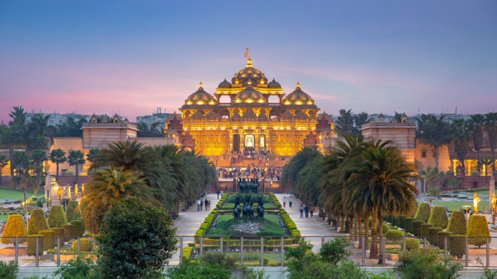 A large, ornate temple, brightly lit, viewed from a distance with symmetrical gardens and palm trees in the foreground during sunset.