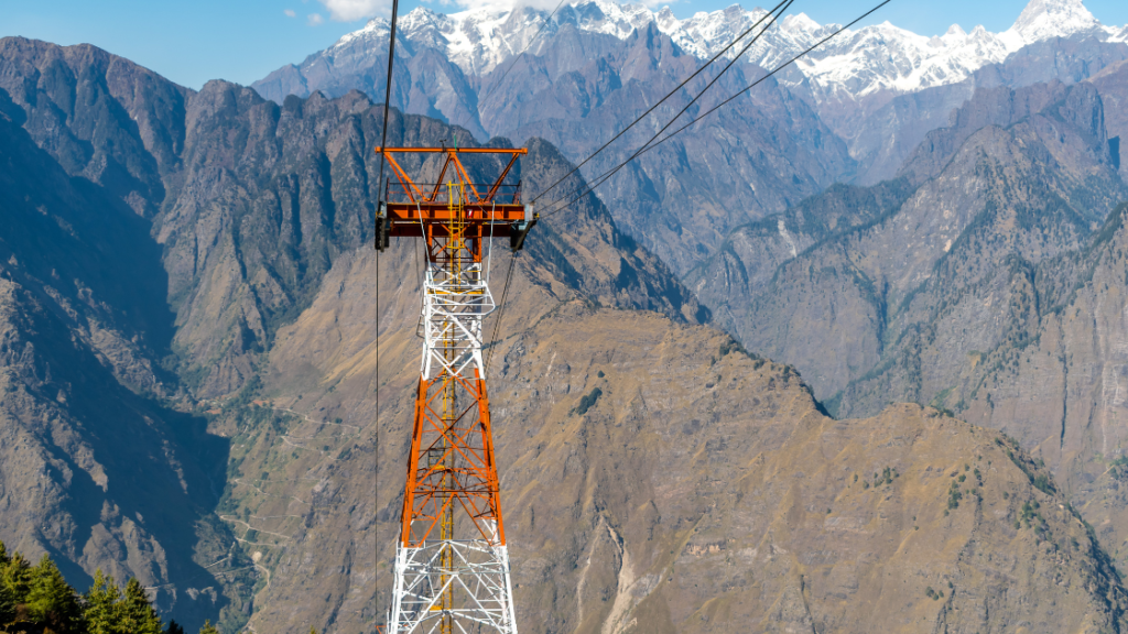 A red and white cable car tower stands amidst a rugged mountainous landscape, with snow-capped peaks in the background.