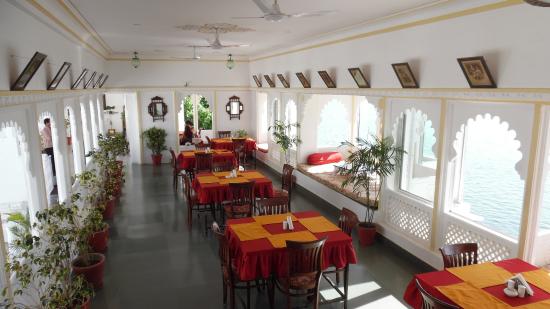 Interior of a bright, airy restaurant with red tablecloths, framed pictures on the walls, and arched windows overlooking water.