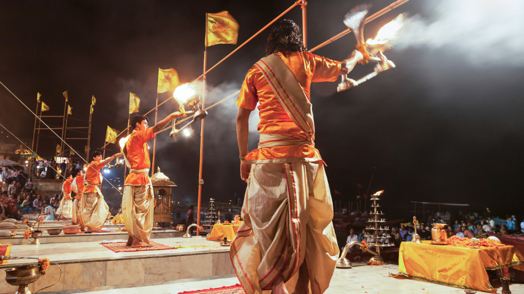 People dressed in traditional attire perform a ritual ceremony involving fire on a platform with yellow flags, witnessed by numerous onlookers at night.