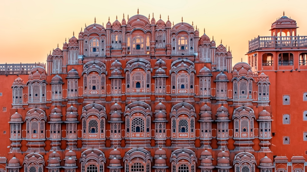 A symmetrical, ornate red and white building with many latticed windows and domes, set against a warm, pastel sky.