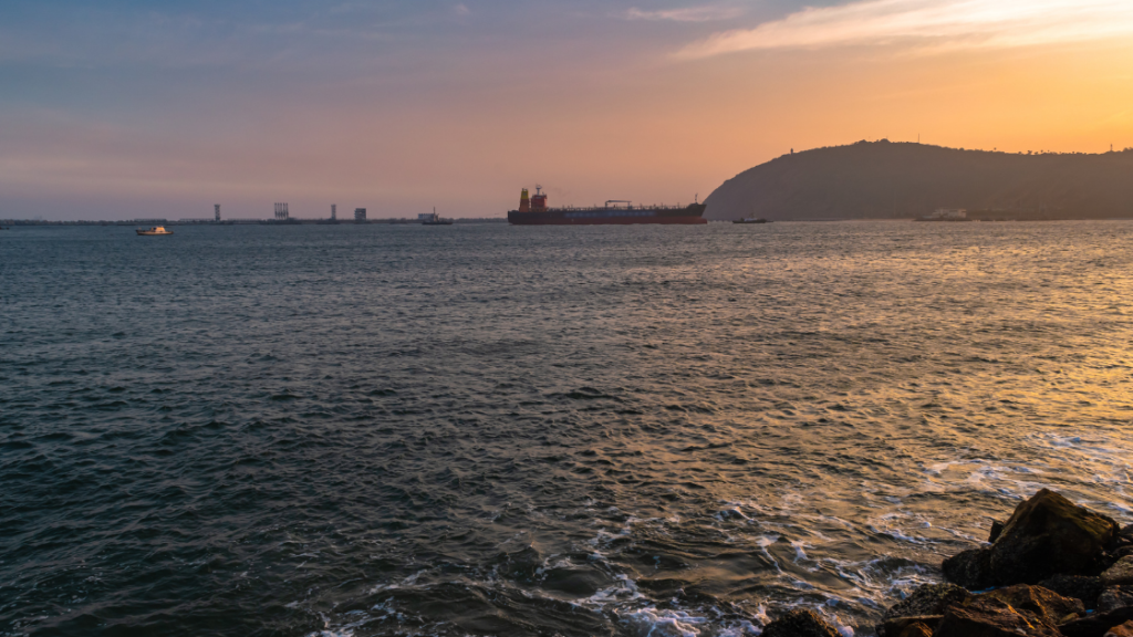 A large cargo ship floats in a calm sea near a rocky shore at sunset, with an industrial skyline and a hill in the background.