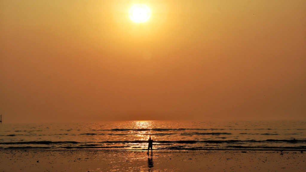 A person stands at the edge of the ocean during sunset, with the sun casting a golden glow over the water and creating a reflection on the wet sand.