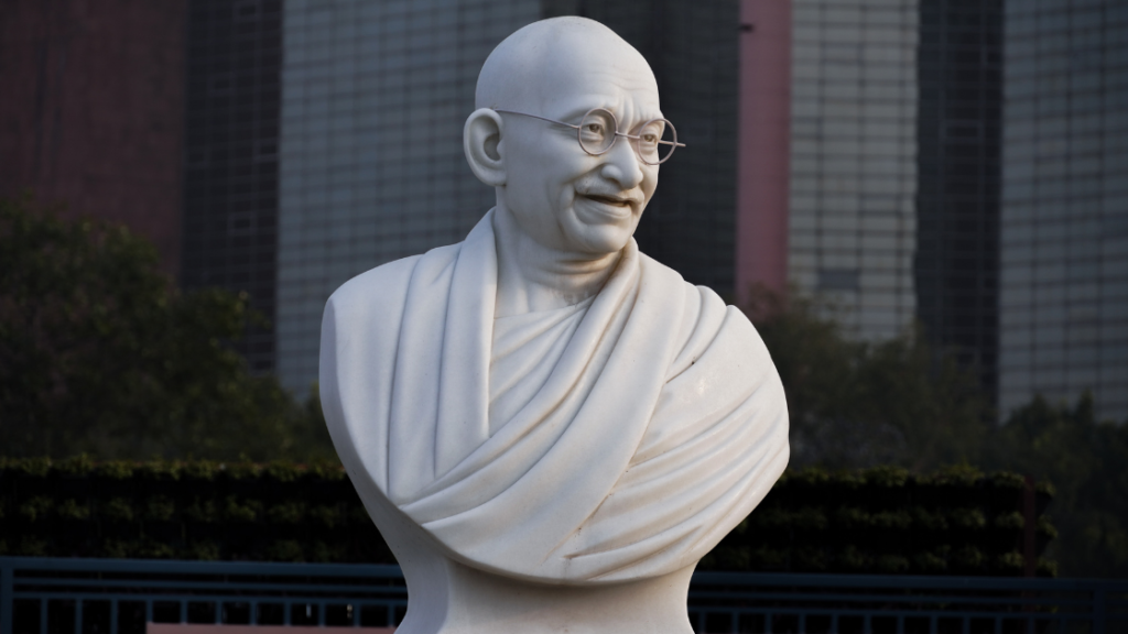 A marble bust of a smiling bald man with glasses and draped garment, set against an urban backdrop with tall buildings and greenery.