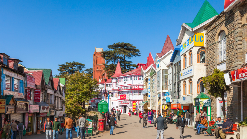 Street scene with people walking past colorful buildings under a clear blue sky. The street includes various shops and businesses. Trees and a red-colored tower are visible in the background.