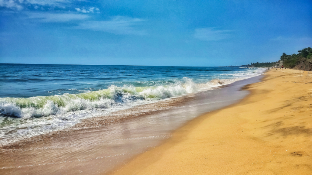 A serene beach scene with golden sand, gentle waves, and a clear blue sky. The shoreline is mostly empty, with some greenery visible in the distance.