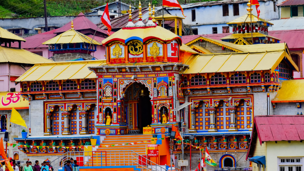 Colorful, intricately designed temple with numerous domes and a central archway. Multiple flags on top, steps and people visible in front. Surrounding buildings have similar architectural style.