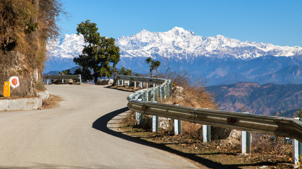 Winding mountain road with guardrails, trees, and a distant snow-capped mountain under a clear blue sky.