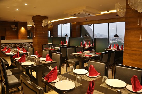 Interior of a modern restaurant with neatly arranged tables, each set with red napkins; large windows offer a view outside.