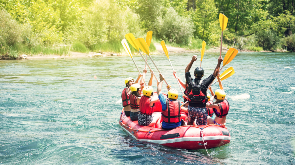 A group of people wearing life jackets and helmets raise their paddles while sitting in a red inflatable raft on a river surrounded by greenery.