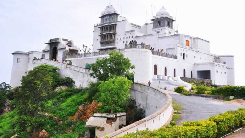 A large white palace with towers and domes sits atop a hill, surrounded by greenery. A winding road leads up to the entrance.