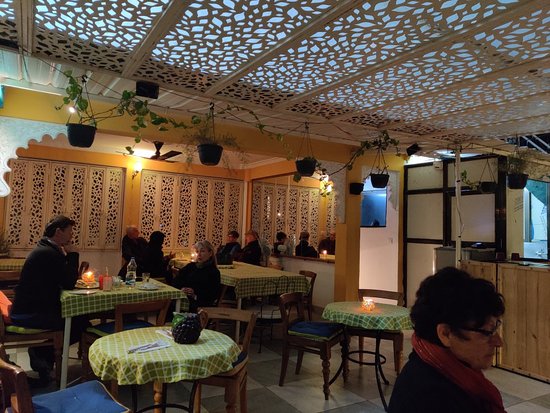 Interior of a cozy café with patterned lattice ceiling, hanging plants, and patrons seated at tables covered with patterned cloths.