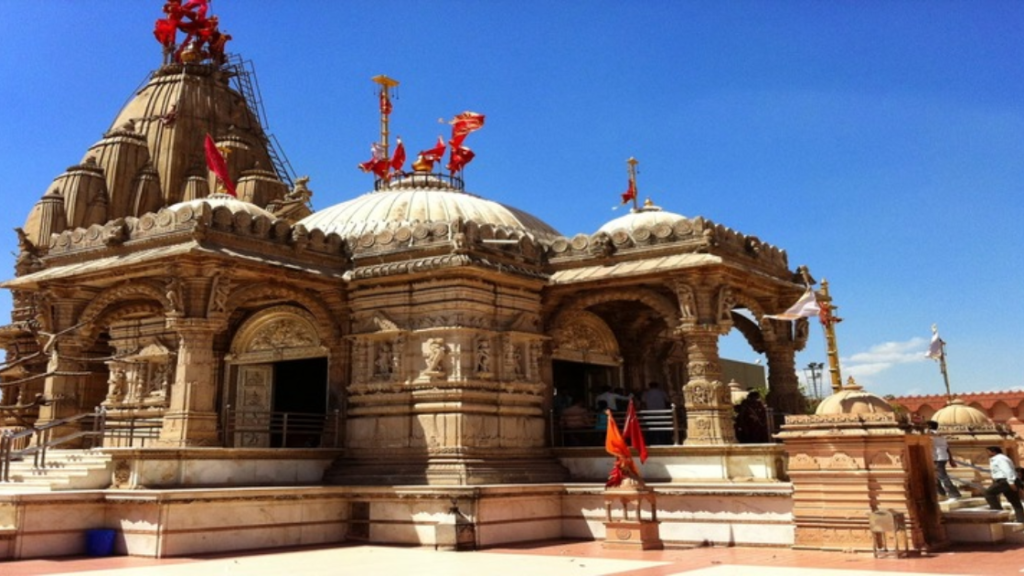 A detailed stone temple with ornate carvings and multiple domes, adorned with red flags, stands under a clear blue sky.