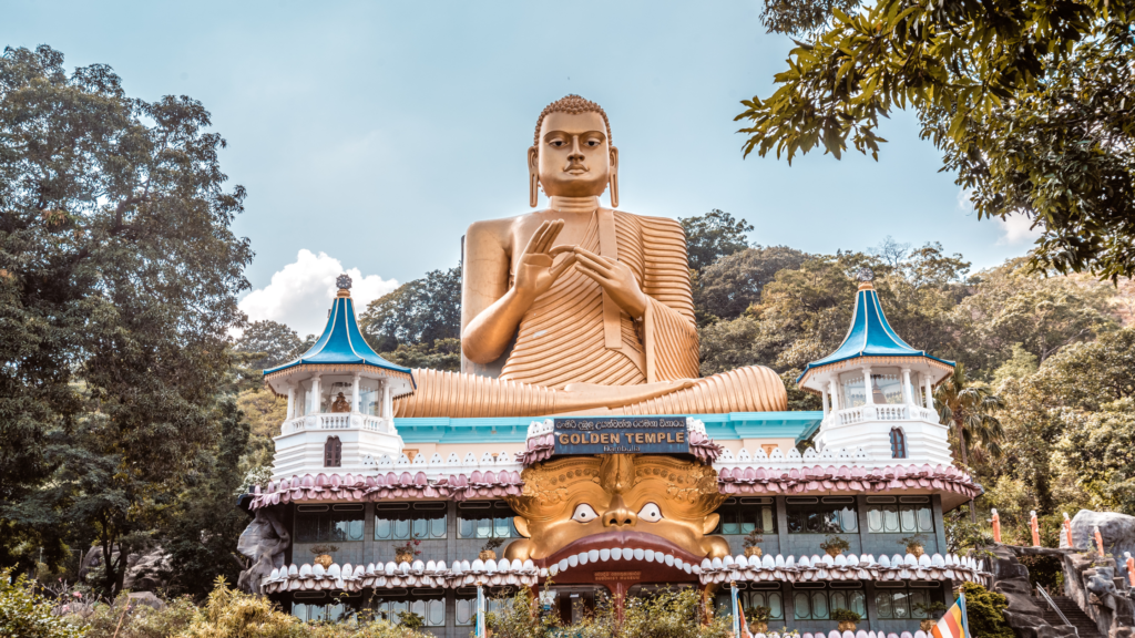 A large golden Buddha statue sits atop a building with a lion's head entrance, surrounded by greenery and trees. The structure has two small towers and is set against a backdrop of hills and blue sky.