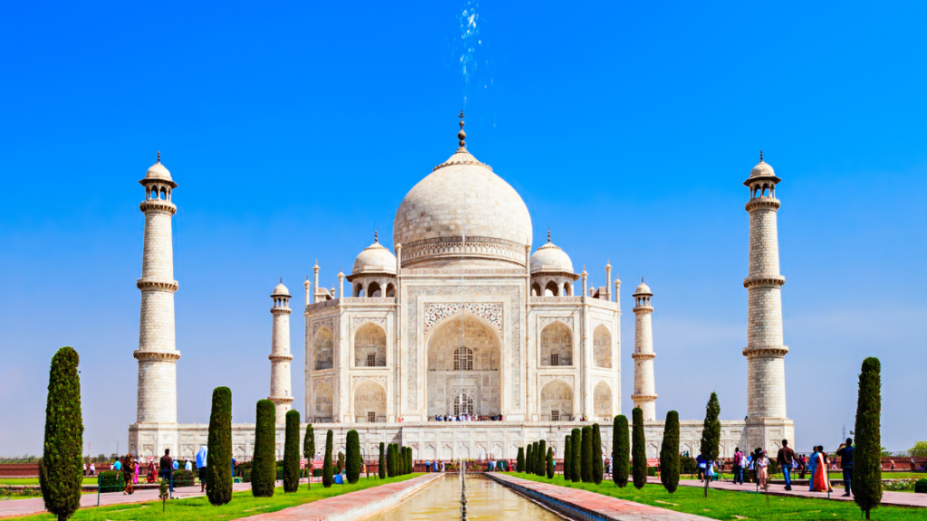 The Taj Mahal, a white marble mausoleum with four minarets and a central dome, stands under a clear blue sky, surrounded by gardens and visitors.