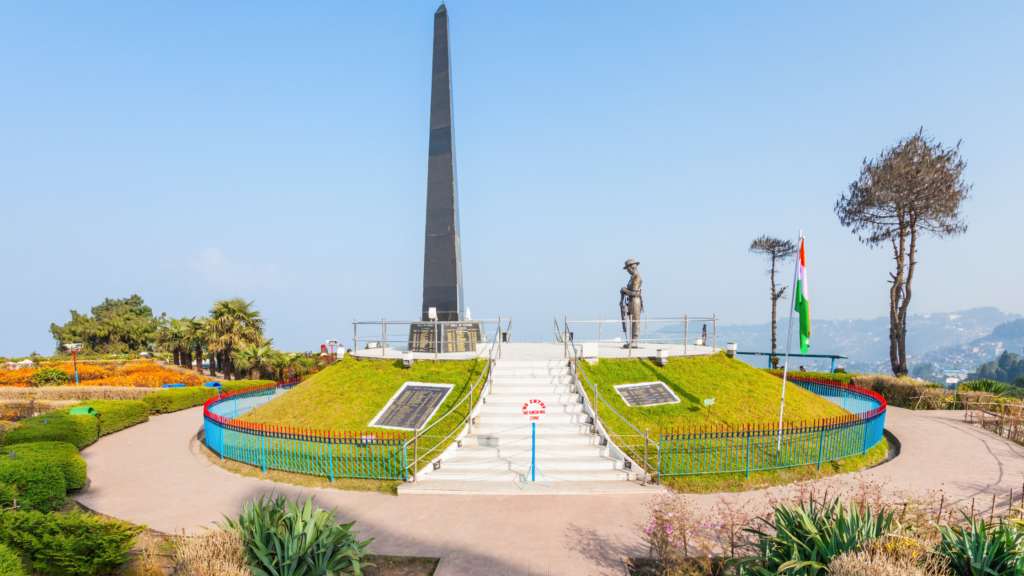 A landscaped garden with a central monument consisting of a tall obelisk, a statue of a soldier, and two plaques, surrounded by green railings and colorful flowers.