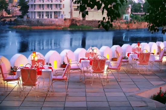 Outdoor dining setup by a riverside at dusk, featuring white chairs, candlelit tables, and arch-shaped white decorations.