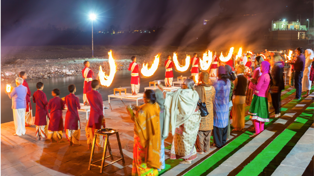 A group of people participate in a night-time religious ceremony near a riverbank, with priests performing rituals involving large fire torches and attendees observing.