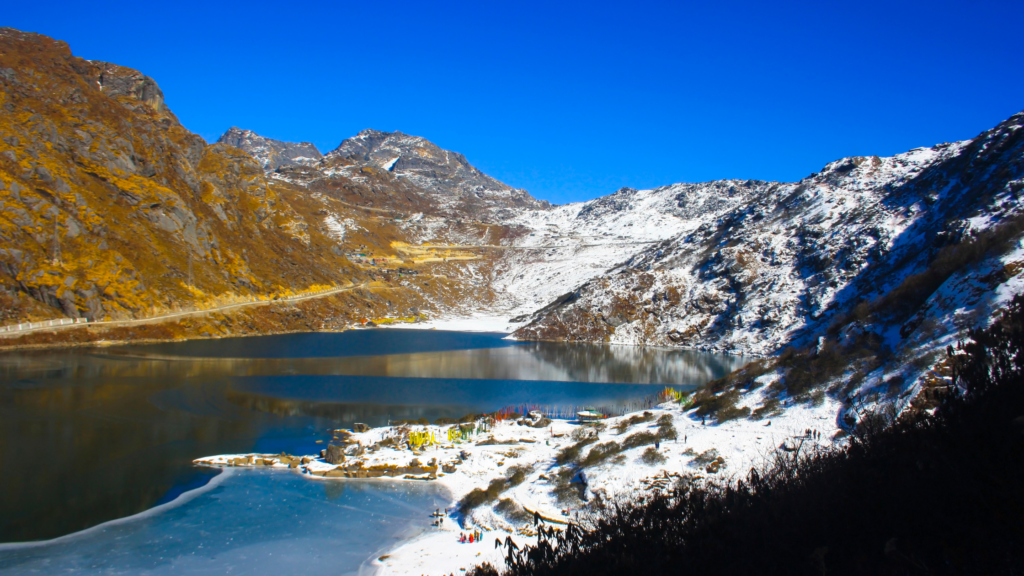 A serene snow-covered landscape featuring a calm lake surrounded by rugged mountains under a clear blue sky. A small settlement is visible near the lake's edge.