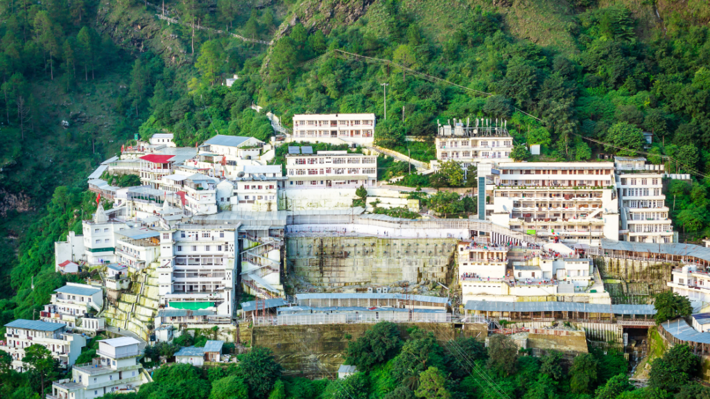 A complex of light-colored buildings is nestled on a hillside covered with dense green trees. The structures are multi-storied and interconnected, with some visible pathways surrounding them.