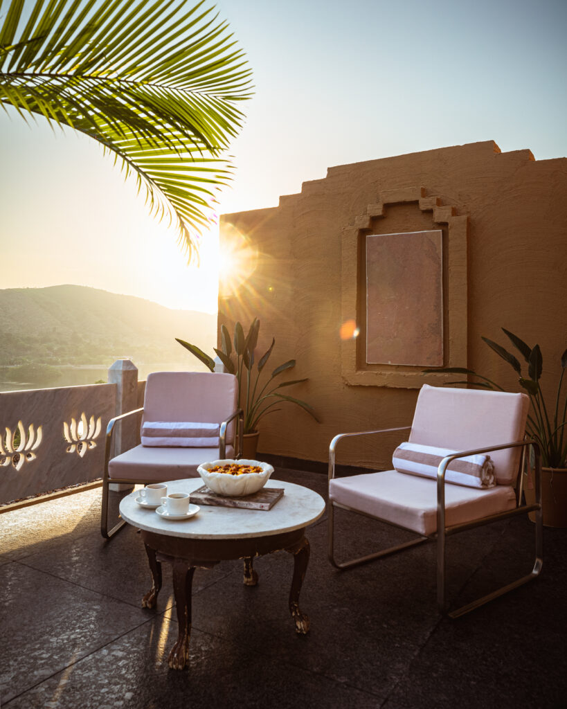 Two cushioned chairs and a small table with tea set on a sunny terrace, surrounded by plants. The sun is setting behind distant hills, casting a warm glow over the scene.