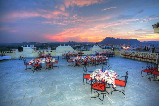 Dusk view from a rooftop restaurant set with tables and chairs, overlooking a scenic landscape with mountains in the distance under a colorful sky.