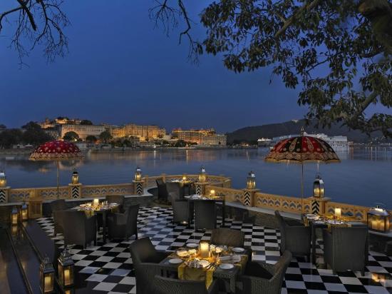 Outdoor dining area with checkered flooring and umbrella-covered tables, overlooking a tranquil lake with illuminated buildings in the background at dusk.