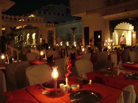 Elegant outdoor dining setup at night featuring tables with red tablecloths and candles, with a traditional white building in the background.