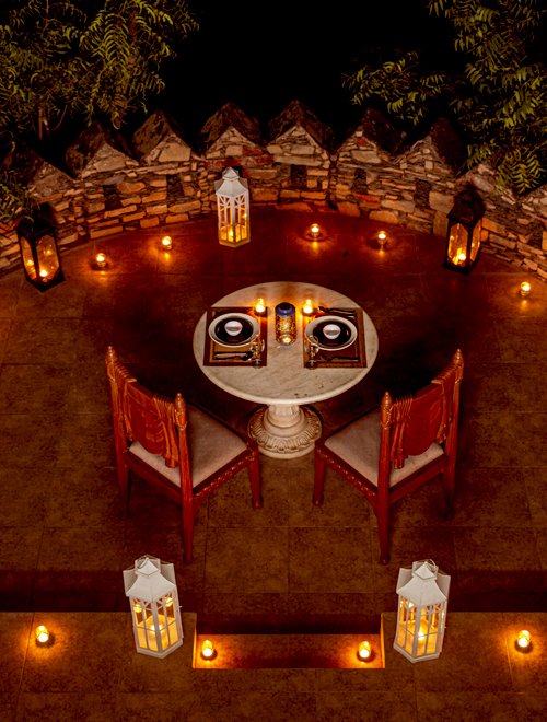 An intimate, candlelit dining setup for two on a patio, nestled within a private pool villa in Udaipur, featuring a round table, two ornate chairs, and lanterns placed around the area for a warm and cozy ambiance.