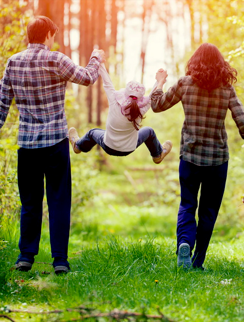 Two adults swing a child by the arms while walking through a sunlit forest, mirroring the joy and togetherness one might experience in a private pool villa in Udaipur. The group appears to be enjoying an outdoor activity together.