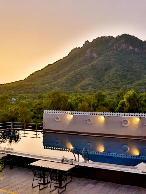 Rooftop pool with lounge chairs and tables, overlooking a lush green mountain range at sunset.