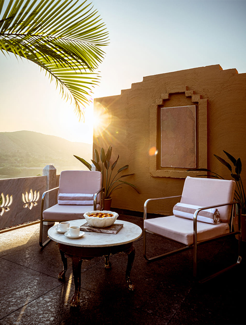 Two chairs with cushions and a small round table with cups and a bowl are on a sunlit patio with a palm leaf and a stucco wall in the background.