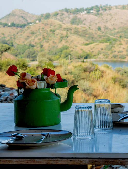 A green teapot with red and peach roses on a table set with plates and drinking glasses, with a scenic hillside and lake in the background.