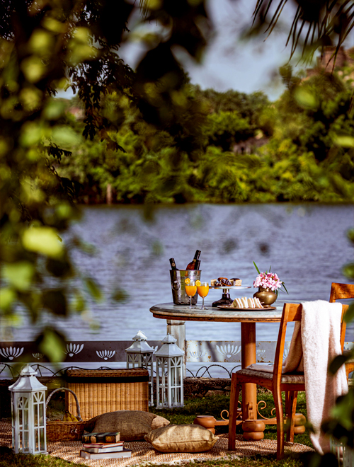 A riverside picnic setup with a wooden table and chairs, featuring various drinks, pastries, and a flower vase. Lanterns, cushions, and a picnic basket are placed around the seating area of your private pool villa in Udaipur.