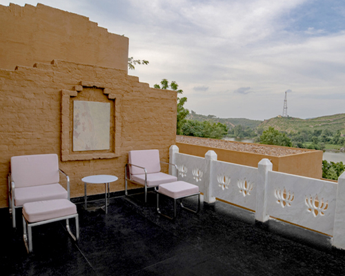 Outdoor terrace with two pink cushioned chairs, a small white table, and a yellow wall with framed artwork. White railing with decorative cutouts runs along the edge. Greenery and hills are visible.