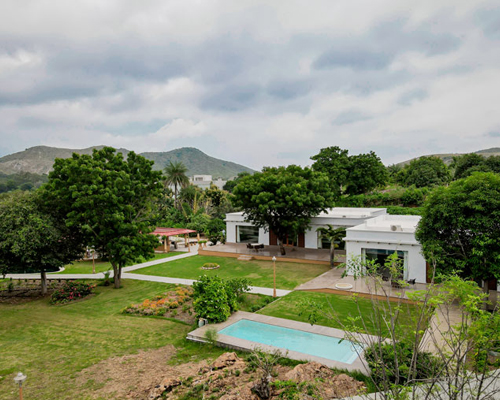 A modern white building with a flat roof is surrounded by a lush green landscape, including trees, bushes, and a trimmed lawn. In the foreground, there is a narrow swimming pool. Hills are visible in the background.