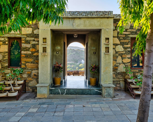 Stone building entrance with potted plants on both sides, leading to a patio with a scenic view of hills and trees in the background.