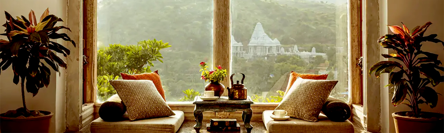 Two cushioned chairs and a small table with a teapot sit by a window framing a scenic view of hills and a distant temple—a perfect setting for your next reflective blog post.