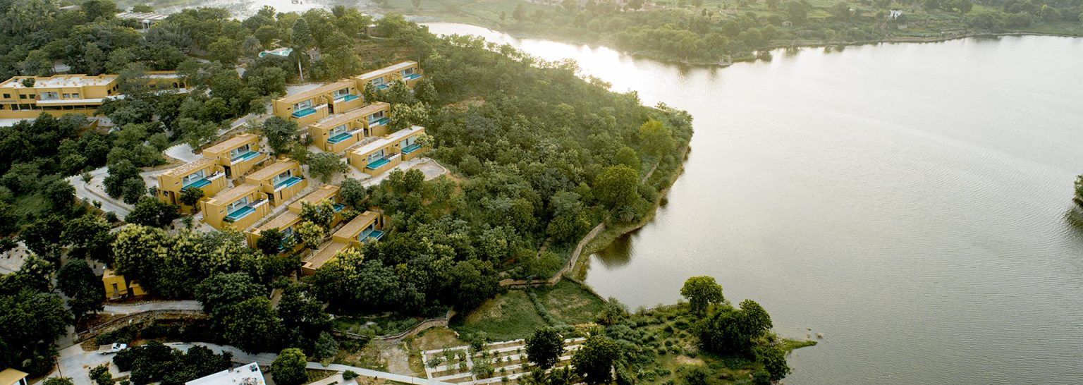 Aerial view of a tranquil lakeside resort with several yellow buildings amidst greenery. The resort overlooks a large, calm body of water on a bright, sunny day.