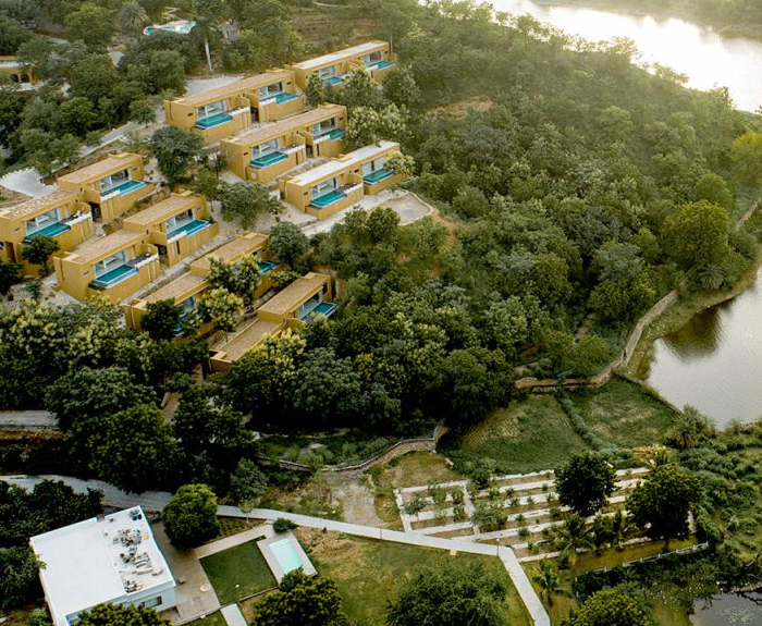 Aerial view of a series of modern villas with private pools nestled in a lush, green landscape near a river. Steps and pathways are visible leading through the surroundings.