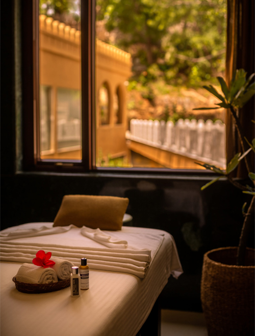 A massage table with neatly folded towels, a red flower, and spa products is situated near a window with a scenic outdoor view.