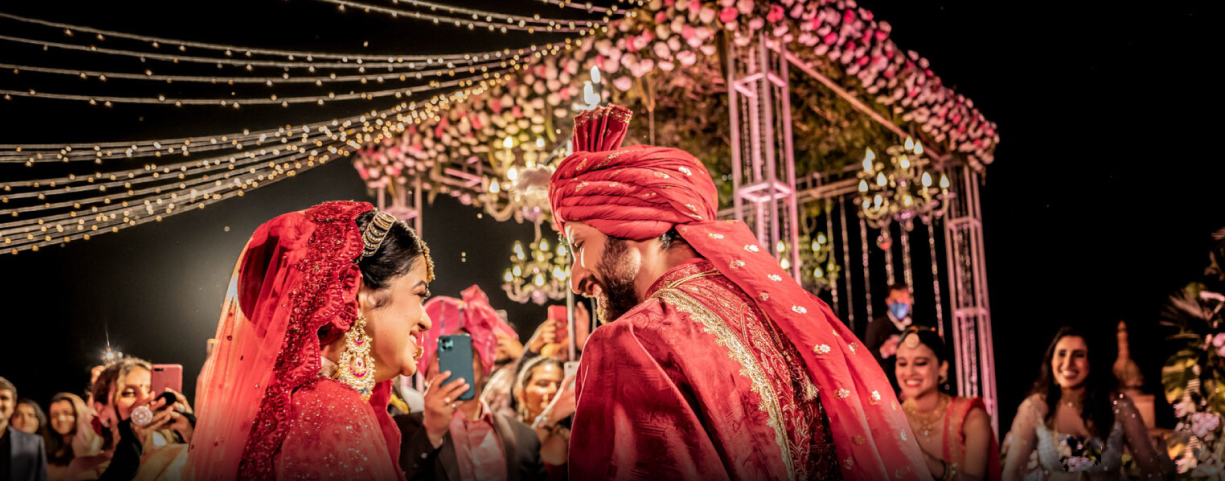 A bride and groom in traditional attire smile at each other under a floral canopy with string lights, surrounded by guests during an enchanting ceremony at a private pool villa in Udaipur.