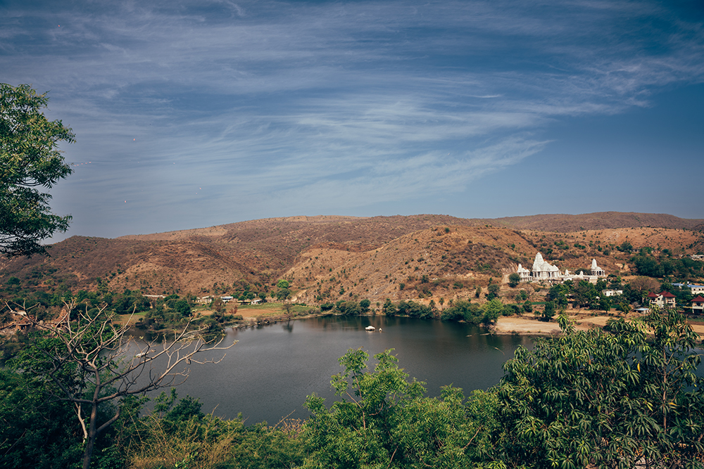 A serene lake surrounded by hills offers the perfect backdrop for a destination wedding, with a white building on the right under a clear blue sky.