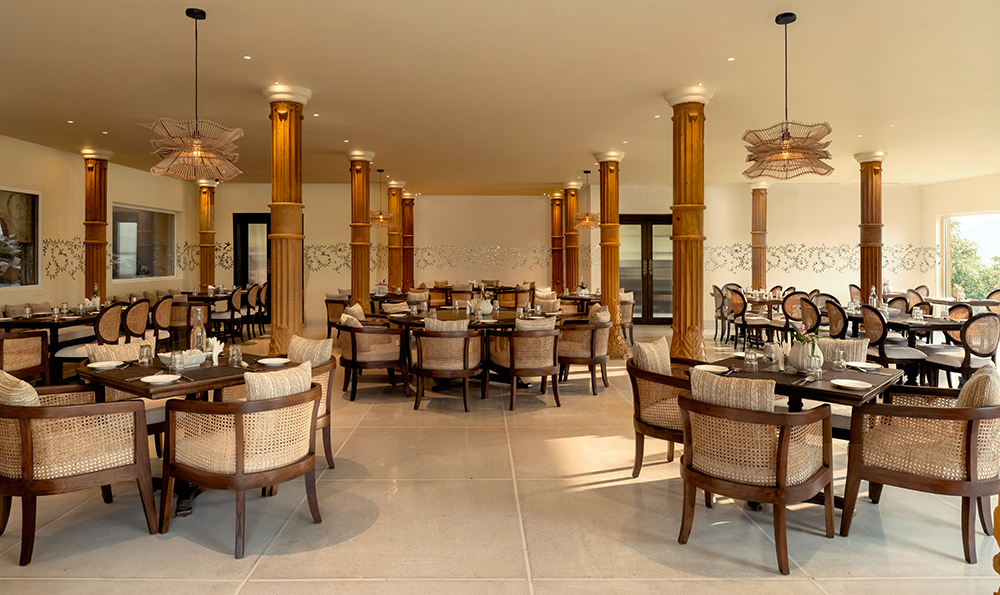 Restaurant interior with wooden columns, wicker chairs, and neatly set tables. Pendant lights hang from the ceiling, and natural light filters in from large windows.