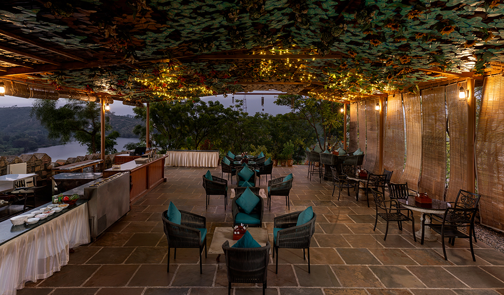 Outdoor dining area with chairs and tables under a leafy canopy. A view of trees and a body of water is visible in the background.