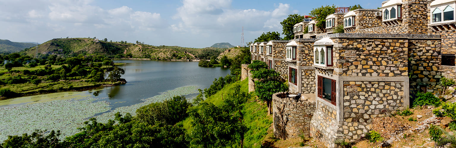 Stone buildings overlook a lush landscape with a lake and hills under a partly cloudy sky.